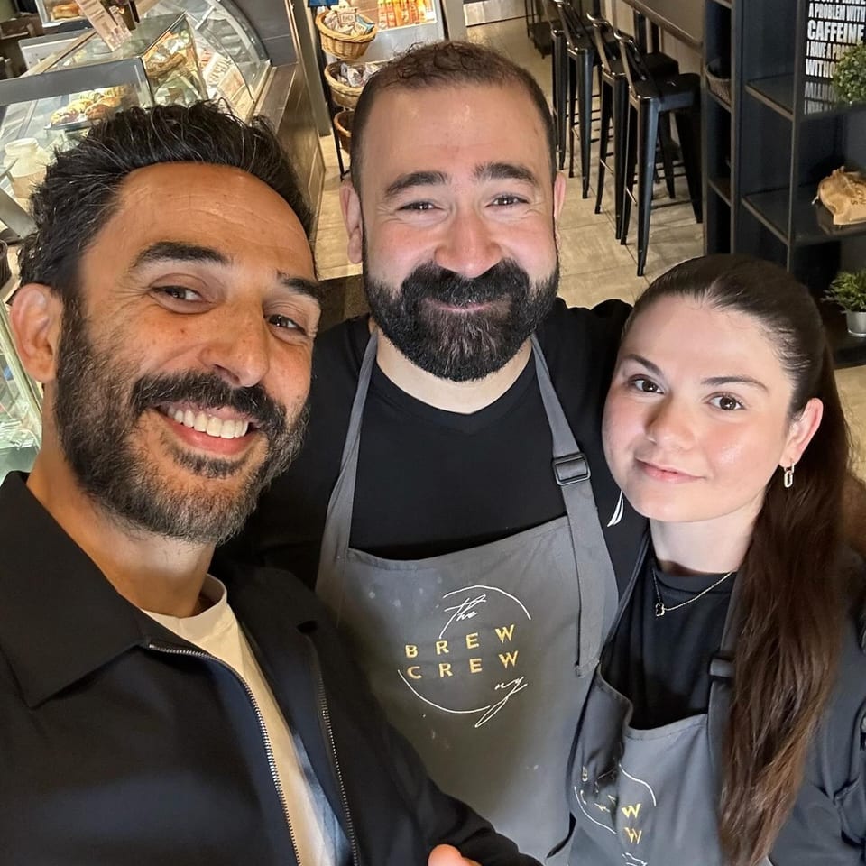 Three smiling people, two wearing "The Brew Crew NY" aprons, posing inside a coffee shop.