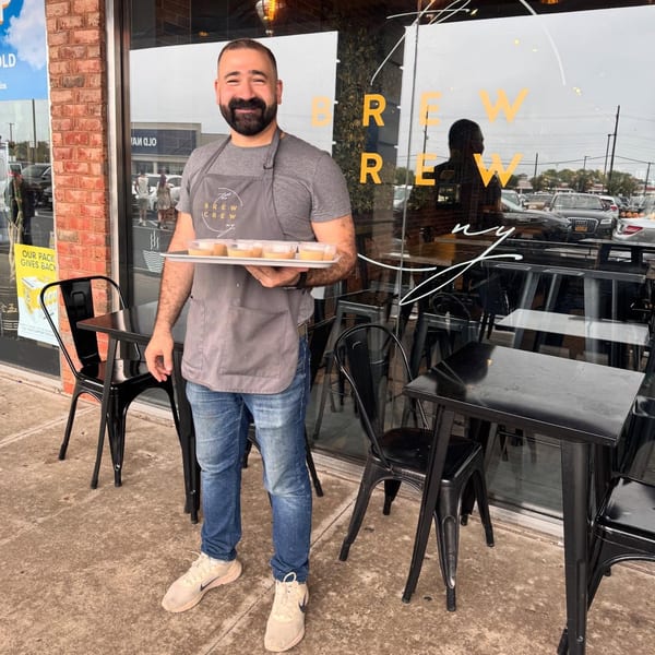 A smiling Brew Crew NY employee in an apron holds a tray of coffee drinks outside the cafe.