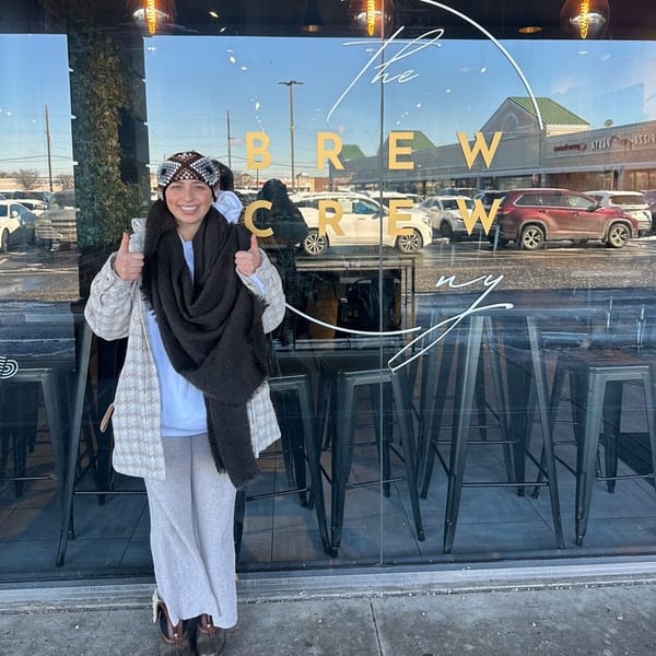 A smiling woman in winter attire gives two thumbs up outside The Brew Crew NY coffee shop.