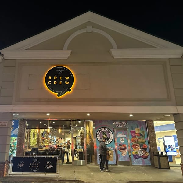 The Brew Crew NY coffee shop exterior at night, with a glowing circular sign and a person entering.